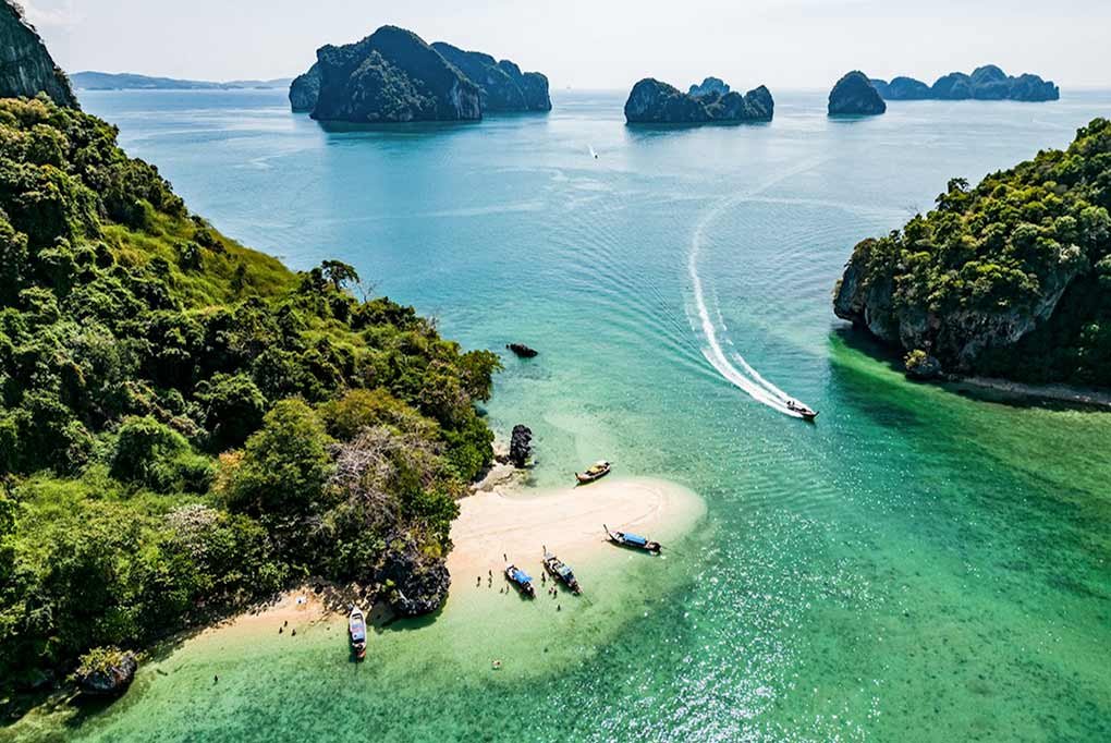 aerial drone shot of phang nga bay limestone islands with a beach in the foreground with traditional thai longtail boats beached with visitors and two more travelling at speed in the channel