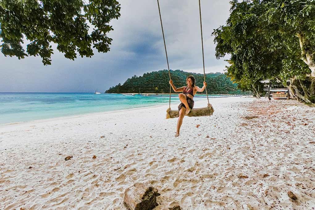 girl on a rope and timber swing beneath a tree on a deserted beach with chalet and small jetty in the background along with 55ft nyami charter catamaran in the distance