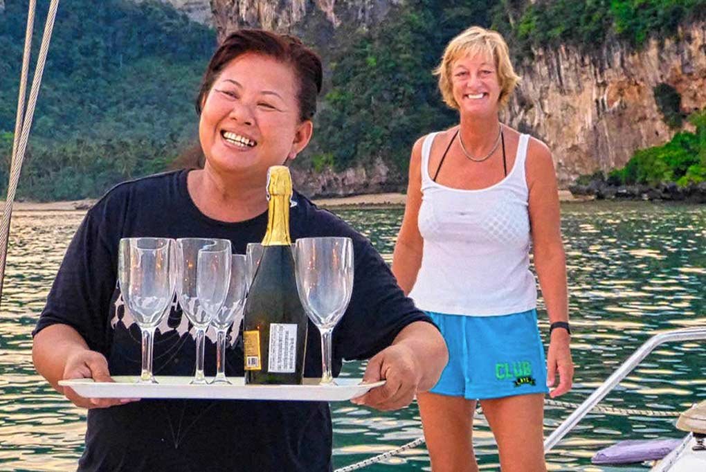 thai staff serving prosecco on deck with nyami at anchor with the backdrop of limestone cliffs and fertile jungle and a very happy looking boat owner following behind the drinks
