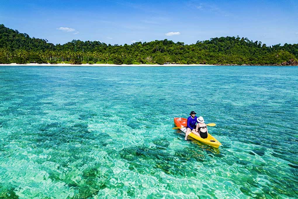 two guests on a red and yellow kayak in shallow tropical waters approacning a deserted paradise beach from their charter catamaran